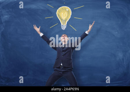 Geschäftsmann mit Händen in den Sieg auf der blauen Tafel Hintergrund angehoben und eine Kreidezeichnung von einer Lampe direkt über ihm. Stockfoto