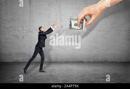 Kleine Geschäftsmann auf konkreten Hintergrund, die heraus für eine riesige Hand Geld. Stockfoto