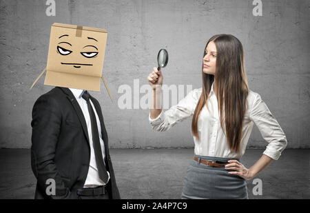 Die Frau ist auf der Suche durch ein Vergrößerungsglas auf ein Geschäftsmann mit einer Box auf seinem Kopf mit einem Pokerface Stockfoto