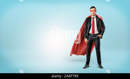 Eine selbstbewusste Unternehmer in einen roten Umhang und eine Maske mit einer Hand in seine Tasche auf blauem Hintergrund. Stockfoto