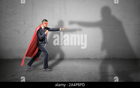 Ein Geschäftsmann in einen roten Umhang und eine Maske aus der Kampf gegen einen Schatten eines anderen größeren Mann auf konkreten Hintergrund. Stockfoto
