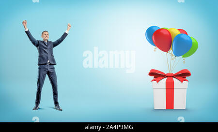 Ein glückliches Geschäftsmann mit Händen erweckt, hinter einem Geschenk Box zu viele Ballons gebunden. Stockfoto