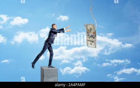 Ein kleiner Geschäftsmann, stehend auf einem konkreten Spalte und fangen einen Dollar Bill auf einer metallenen Haken verfangen. Stockfoto