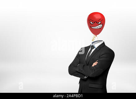 Ein Geschäftsmann auf weißem Hintergrund steht mit gekreuzten Händen und einem roten wütend das Gesicht Ballon anstelle von seinem Kopf. Stockfoto