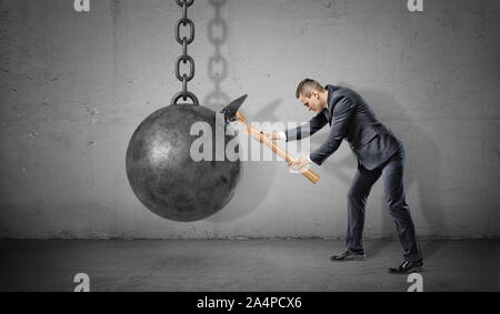 Ein kleiner Geschäftsmann hits einer großen Festen eisernen Kugel mit einem Brechen der Hammer auf konkreten Hintergrund. Stockfoto