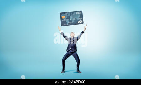 Ein glückliches und siegreichen Geschäftsmann steht auf einem blauen Hintergrund und hält ein riesiges generische Kreditkarte. Stockfoto