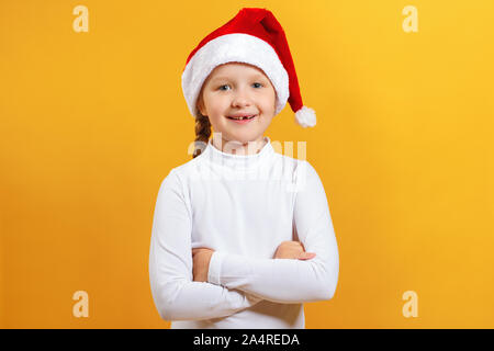 Charmantes Portrait Of Happy süsse kleine Mädchen in Santa hat. Das Kind, verschränkte die Arme und schaut in die Kamera auf einem gelben Hintergrund. Stockfoto