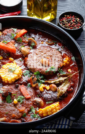 Nahaufnahme der Hackbraten Burger geschmort in marinara Sauce, mit Pilzen, Gemüse und Hühneraugen in einem schwarzen Keramik Schmortopf mit Zutaten auf einem schwarzen Sto Stockfoto