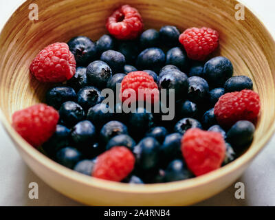 Heidelbeeren und Himbeeren in eine hölzerne Schüssel auf weißem Marmor Hintergrund im Studio. Stockfoto