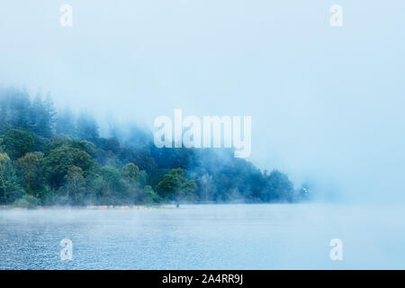 Dawn Cloud beginnt, scheint die Sonne durch den Nebel über einem Waldgebiet am See Bassenthwaite Lake District, Cumbria, England zu zerstreuen. Stockfoto