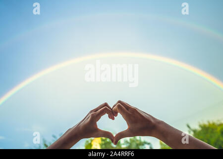 Herz aus Hände auf einem Regenbogen Hintergrund. LGBT-Flagge in der Form eines Herzens gemalt. Symbolik der Welt. Stockfoto