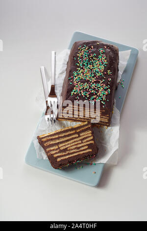 Blick von oben auf die Schokolade Kuchen mit Keksen und bunten Topping, serviert mit zwei Gabeln auf blau Keramik Schale und weißes Papier. Traditionelle ungekocht Kalter Stockfoto