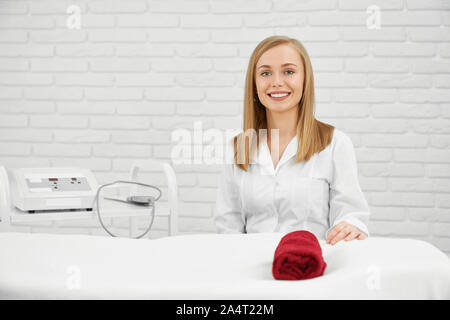 Positive, junge Frau im weißen Kittel massage Tisch sitzen, von der Kamera suchen, lächelnd. Porträt der jungen weiblichen professionellen Kosmetiker. Konzept Stockfoto