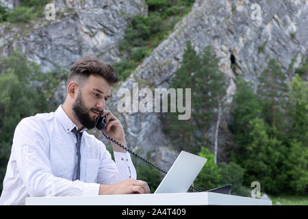 Arbeitnehmer Mann an Schreibtisch mit Computer Laptop und Telefon am Arbeitsplatz, an Bildschirm schaut. Outdoor junge Hipster in Gespräch am Telefon Stockfoto