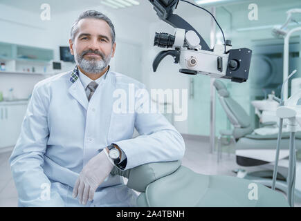 Stattliche Zahnarzt in Stuhl, posierend, an der Kamera schaut. Reifer Mann im weißen Kittel in private Zahnheilkunde Klinik mit moderner Ausrüstung, professionelle Mikroskop Bärtigen. Stockfoto