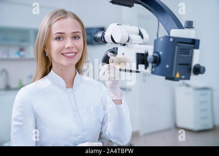 Adorable blonde Frau arbeiten als stomatologist in private Zahnheilkunde Klinik. Fröhliche Zahnarzt an der Kamera suchen, lächeln und posieren. Spezialisten arbeiten mit moderner Ausstattung wie zahnmedizinische Mikroskop. Stockfoto