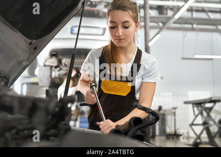 Charmante, hübsche junge Frau arbeiten und Reparatur Auto Autoservice. Mechanikerin holding Spezialwerkzeug, Reparatur Auto unter der Motorhaube. Mädchen tragen in Overalls, weißes T-Shirt. Stockfoto