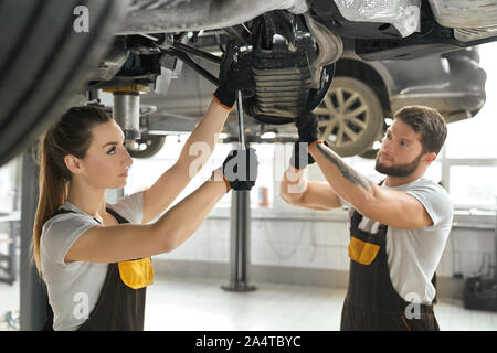 Junge Frau und Mann gemeinsam mit Fahrwerk des Automobils. Mechanik tragen in den weißen T-Shirts und Overalls. Arbeitnehmer mit Werkzeugen, Schlüsseln für die Festsetzung Fahrzeug in Autoservice. Stockfoto