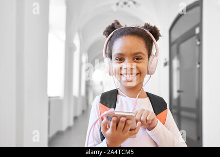 Schön, positive Schulmädchen mit Kopfhörern holding Telefon, posierend, an der Kamera schaut. Glücklich, schönes Kind stehen in langen Flur, hören Musik und lächelnd. Stockfoto