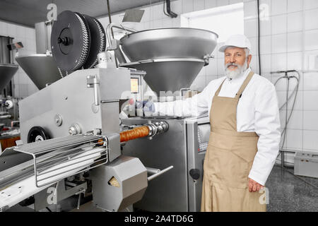 Ältere Menschen arbeiten auf der Wurstherstellung. Professionelle Arbeiter in Uniform tragen, stehen in der Nähe von Anlagen, Posieren und Kamera. Metzger in der modernen Fabrik arbeiten. Stockfoto