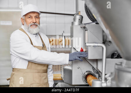 Metzger Mann arbeiten an Wurst und Fleisch Produktion. Professionelle Mitarbeiter in weiße Uniform in der Nähe von neue, moderne Ausrüstung, posing und Kamera. Stockfoto