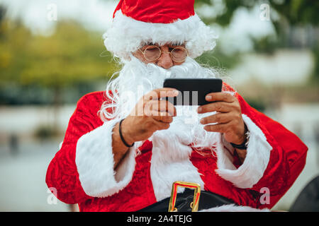 Foto von Santa Claus in einem Gespräch mit einem Mobiltelefon mit einem Park hinter unfocused. Weihnachten Stockfoto
