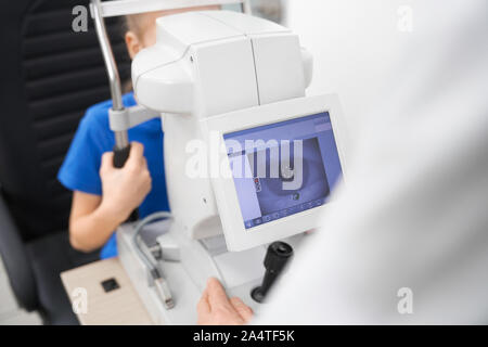 Selektiver Fokus der Maschine für die Überprüfung des Sehvermögens in der Klinik. Männlicher Arzt mittels Spaltlampe und der Prüfung Auge Pupille der kleinen Patienten im Labor. Konzept der Vision Korrektur und Gesundheit. Stockfoto