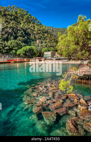 Herrlicher Blick auf die Bucht Paraggi in Italien, mit Steinen in klares Wasser im Vordergrund, Hügel mit grünen Bäumen, weit entfernten Strand und Pier abgedeckt, Schuß auf su Stockfoto