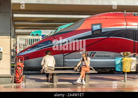 Ein Mädchen sitzen auf einer Bank im Gespräch über ihr Mobiltelefon vor einem Trenitalia Weingut Frecciarossa 1000 High Speed Zug am Bahnhof Porta Nuova, Turin Stockfoto