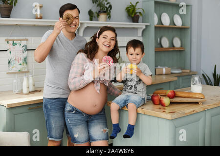 Glückliche junge Familie, eine schöne schwangere Frau mit ihrem stattlichen Mann und Baby boy Frühstück machen und Essen lecker Donuts in der Küche in der Stockfoto