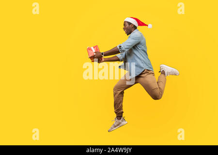 Volle Länge Seitenansicht des freudigen hübscher junger Mann in Santa Hut und casual denim Shirt springen oder Laufen mit Weihnachten Geschenk Box, Eile für den Vertrieb. Stockfoto