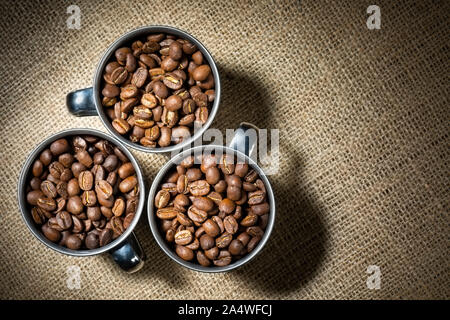 Drei Tassen Kaffee Bohnen auf einem hessischen Hintergrund mit Kopie Raum auf der rechten Seite. Das Metall Becher sind in einem Kreis unter einem hellen Licht angeordnet. Stockfoto