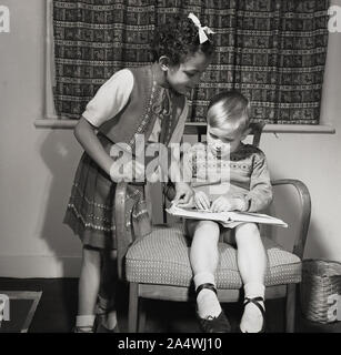 1960er Jahre, historisch, ein junges karibisches Mädchen mit Haarschleife, neben einem kleinen Jungen, der auf einem Stuhl sitzt und ihm beim Lesen seines Buches hilft. Stockfoto