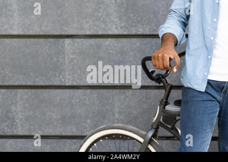 Nicht erkennbare schwarze Mann stand in der Nähe von Fahrrad gegen graue Wand Hintergrund Stockfoto