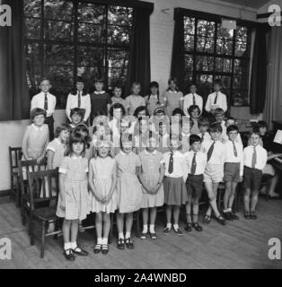 1967, historisch, eine Gruppe uniformierter Grundschulkinder in einem Schulchor, die vor ihren Stühlen in einem Saal stehen, mit ihrem Musiklehrer neben ihnen, am Klavier sitzen, England, Großbritannien. Stockfoto