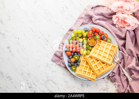 Ein Teller mit frischem Obst und Waffeln. Die Früchte sind Mini Kiwi, Erdbeeren, Passionsfrucht, Weintrauben, Himbeeren und Heidelbeeren. Mit einem purpl Stockfoto