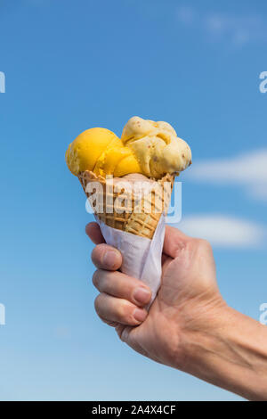 Ein Eis Waffel Kegel mit einem blauen Himmel im Hintergrund. Gelb, beige und braun gelato Eis in ein Eis Waffel Kegel, mit einem blauen Himmel im Hintergrund. Stockfoto