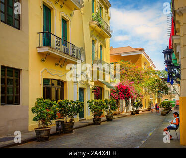 Havanna, Kuba, Juli 2019, Blick auf eine Frau sitzen und lesen in der Calle Mercaderes eine gepflasterte Straße voller Pflanzen in Töpfen und blühende Bäume Stockfoto