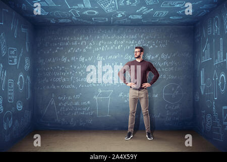 Ein Mann in Gläsern steht in einem kleinen quadratischen Raum, wo die Wände mit Kreide Gleichungen und Diagrammen abgedeckt sind. Stockfoto