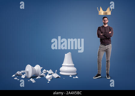 Ein bärtiger Mann steht mit einer großen goldenen Krone über den Kopf und in der Nähe eines großen gebrochen Schach Abbildung. Stockfoto