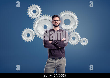 Eine Smart Casual Mann trägt eine Brille und steht in einer Vorderansicht mit mehreren großen Zahnräder hinter ihn anspornen. Stockfoto
