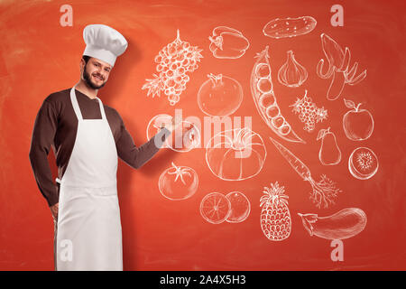 Eine nette junge Küchenchef in einem Chef hat und Schürze, eine Präsentation Geste auf ein Bild von Obst und Gemüse auf eine Wand. Stockfoto