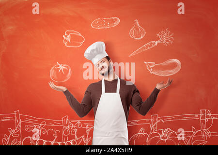 Eine nette junge Küchenchef in einem Chef hat und Schürze, eine Präsentation Geste auf Bilder von Gemüse auf eine Wand. Stockfoto