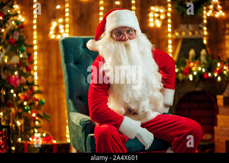 Foto von Santa Claus in Brillen sitzen auf einem Stuhl und schaut an Kamera Stockfoto
