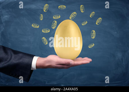 Ein Geschäftsmann hand mit einem großen goldenen Ei auf einem blauen Hintergrund mit Blackboard gezeichnet Dollar Münzen. Stockfoto