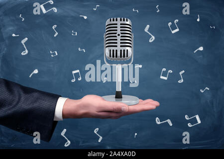 Eine kleine silberne retro Mikrofon stehend auf der Hand eines Kaufmanns unter Noten auf einer Tafel Hintergrund dargestellt. Stockfoto