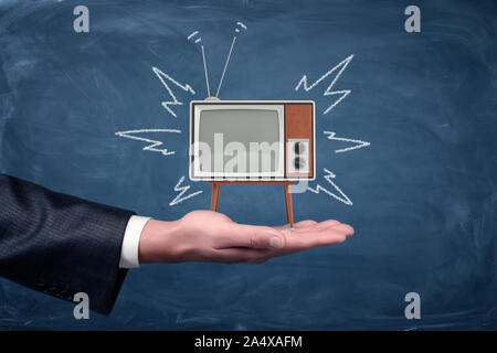 Ein Geschäftsmann Palm hält eine kleine retro TV mit Beinen und Antenne auf blackboard Hintergrund. Stockfoto