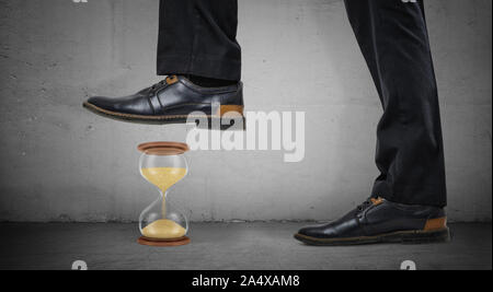 Riesige männliche Beine in glänzenden Stiefel stehen auf konkreten Hintergrund und Schritt auf eine kleine Sanduhr. Stockfoto