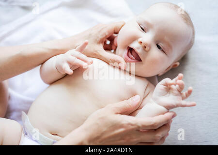 Fröhliche niedlichen Baby Mädchen liegt auf dem Bett, während Mutter bei Ihrem ersten Zähne suchen Stockfoto