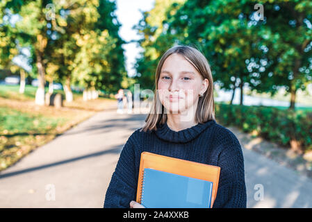 Mädchen teenager Schulmädchen, Sommer Stadt Hintergrund Bäume und Strasse, warmer Pullover, in den Händen von Notebooks Bücher, lächeln, glücklich, nach dem Unterricht in der Schule und Stockfoto